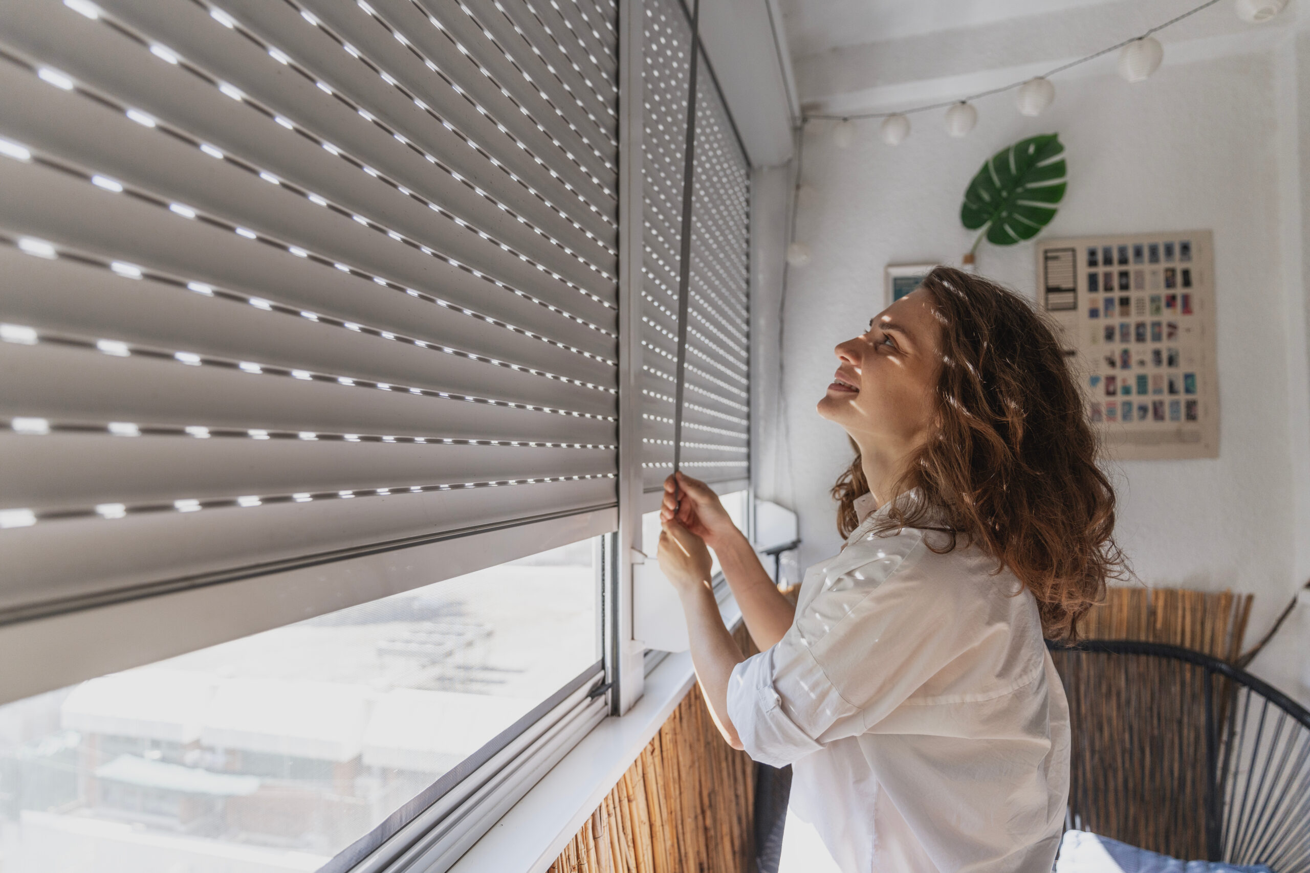 A young woman is opening roller blind shutters on the balcony on a summer day.